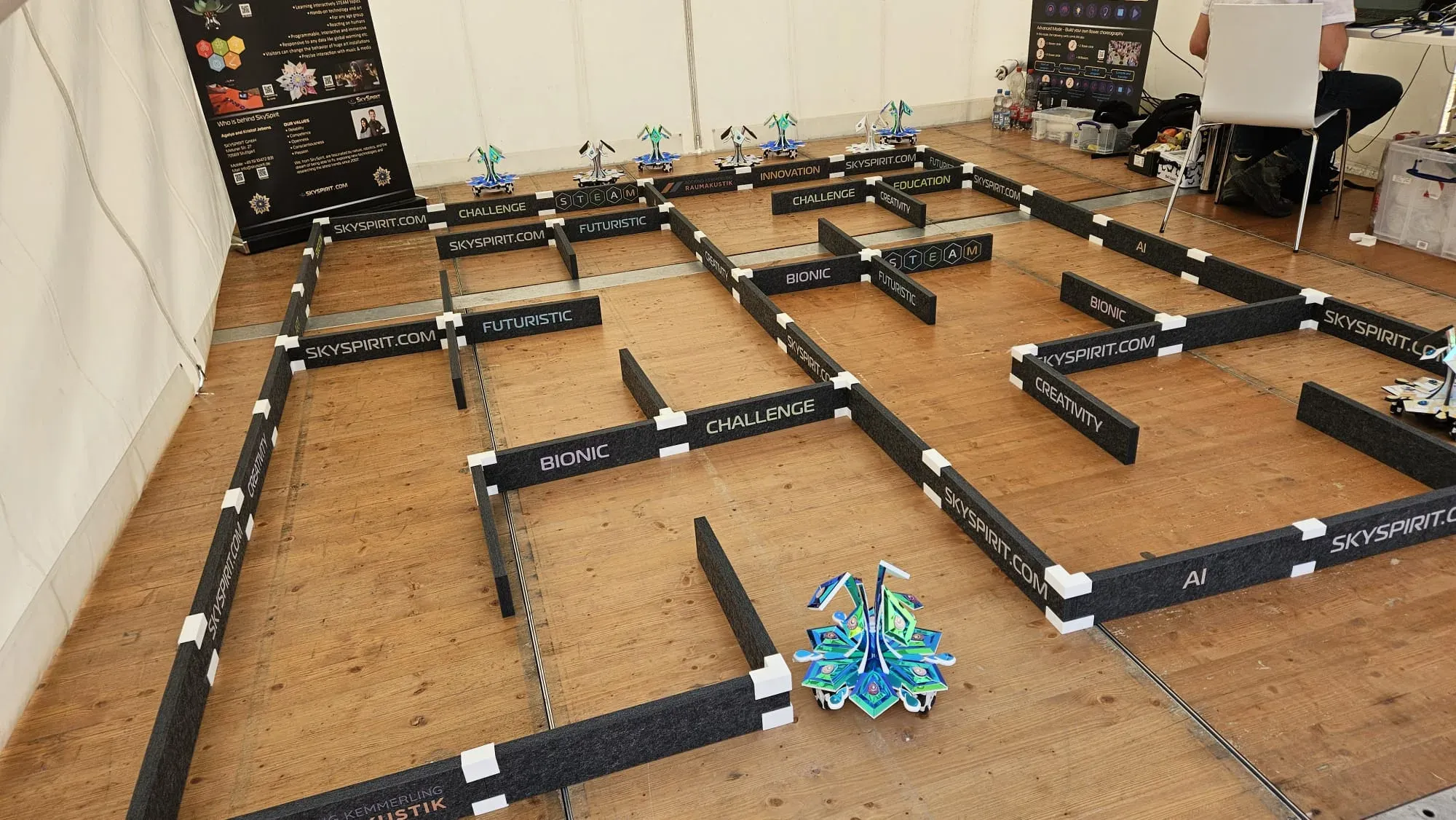 The robot maze in the festival tent: low dark walls on a wooden floor, a flower-shaped robot at the entrance and two SkySpirit roll-up banners behind